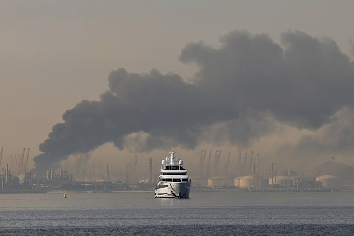 A yacht sails on the water as thick black smoke rises from the port of Jebel Ali in Dubai, with industrial structures in the background